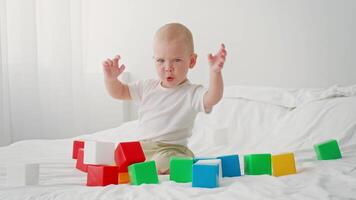 A young child sits on a bed surrounded by colorful building blocks, expressing joy and curiosity. The bright room provides a cheerful atmosphere for playtime and discovery. video