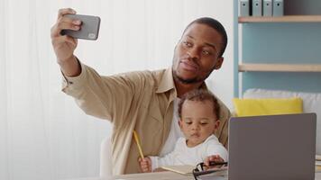 A joyful working father interacts with his toddler indoors, using a smartphone to capture their moment together. The child is sitting on his lap, showing curiosity and excitement. video