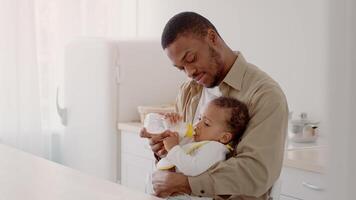 A young African American father lovingly holds his baby while feeding them a bottle. The soft indoor lighting creates a warm and nurturing atmosphere for this intimate moment. video