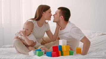 A toddler sits on a bed with parents, sipping from a bottle. Colorful blocks are scattered around as they engage in playful interactions, enjoying a warm, nurturing environment. video
