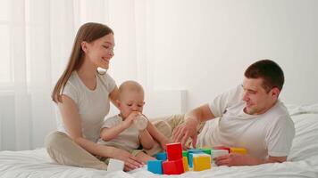 A toddler enjoys playtime on a comfortable bed while parents engage with colorful building blocks. The bright, airy room creates a warm atmosphere for family bonding. video