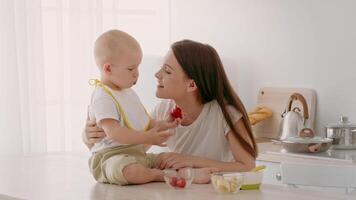 A toddler sits on the kitchen counter sharing a snack with mom. The atmosphere is warm and bright, showcasing a cozy home environment filled with love and care during mealtime. video