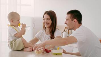 A joyful toddler sips from a bottle while sitting at a table with parents. The family shares snacks and smiles in a modern, sunlit kitchen, enjoying quality time together. video