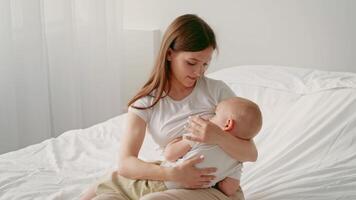 A mother tenderly holds her baby on a bed in a softly lit room. The child seems relaxed and content. This warm moment highlights the bond between them in their comfortable home environment. video