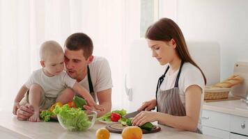 A toddler sits on a countertop while parents chop vegetables and prepare a healthy meal. The bright kitchen, filled with fresh produce, showcases family bonding during meal prep. video