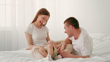 un Pareja se sienta en un suave cama, jugando y riendo con su alegre niño pequeño. el brillante habitación es lleno con natural luz, creando un calentar y amoroso atmósfera para familia vinculación video
