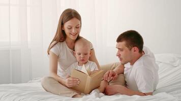 A child sits between parents on a bed while they read together. The family shares smiles in a bright, airy room filled with soft light. It captures a warm, nurturing atmosphere. video