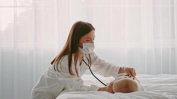 A medical professional, wearing a mask, gently examines a toddler lying on a bed with sunlight streaming through sheer curtains. The setting is a calm home environment focusing on child care. video