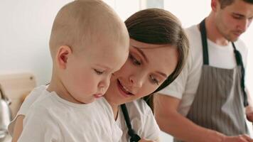 A mother lovingly engages with her toddler in a warm home setting, focusing on the child's curiosity while a father prepares nearby. The atmosphere reflects care and togetherness. video