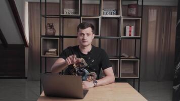 Man working on a laptop in a modern office with bookshelves, gesturing while filming or chatting video