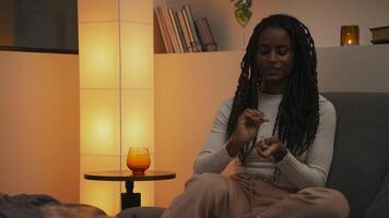 Young African American woman lighting a candle with a match and placing it on a table, then sitting down comfortably and gazing at the flame in a cozy evening setting. video