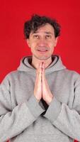 cheerful curly man, dressed in gray hoodie, prays with palms of hands folded in front. Isolated red background of studio shot. Concept - prayer, religion, petition, anticipation video