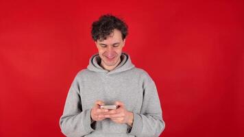 young adult, curly man in gray hoodie, writes text message, comments on mobile phone, texts in messengers. Isolated red background of studio shot. concept - communication, technology, social media. video