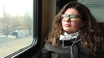 young woman in glasses listens to music through headphones while gazing out train window, capturing serene moment of introspection during urban commute Soft natural light highlights focused expression video