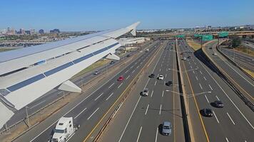 Airplane landing near a city, wing and runway in view with vehicles and buildings around, urban highway running parallel in a clear sunny afternoon video