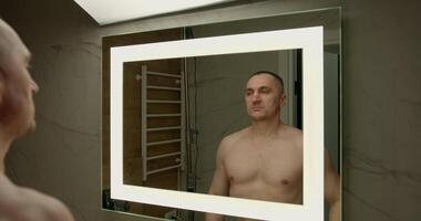 man is standing shirtless in front of a well lit mirror confidently flexing his muscles while examining his physique in the contemporary bathroom setting video