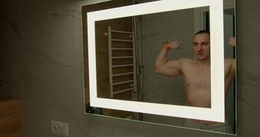 In a modern bathroom illuminated by natural light a man poses flexing his muscles in front of a mirror. He displays a proud expression while enjoying his reflection and fitness journey video