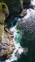 Aerial view of a coastal landscape with cliffs, green fields, two people on the observation deck, and a shimmering ocean under the sunlight with a slow movement up and away video