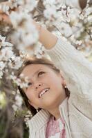 In a serene moment, the girl stands in front of a blooming almond tree, her soft expression captured against the backdrop of vibrant white and pink blossoms photo