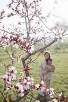 In the heart of spring, a boy and girl stand together under a blooming tree with pink petals, their arms around each other and smiles that light up their faces photo