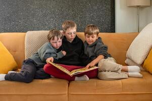 A teenage boy sits in the middle of a couch, reading a storybook to his two younger brothers, who lean in eagerly. photo