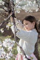 A young girl stands beneath an almond tree in full bloom, surrounded by delicate white petals swaying in the breeze. photo