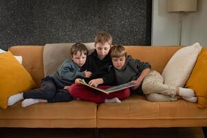 An older boy reads a book to two little boys on the sofa, their faces showing interest and excitement. photo
