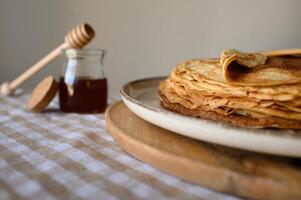 Stack of pancakes with honey on a cozy morning table. A homemade breakfast with golden-brown pancakes, a honey jar, and a honey dipper placed on a softly lit tablecloth. photo