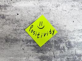 Yellow sticky note in a diamond shape displaying the word Positivity accompanied by a smiley face on a textured grey background, representing the essence of optimistic thinking and cheerful outlook photo