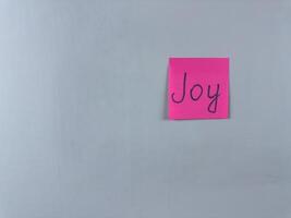 Sticky note with Joy written on it, affixed to a white background. Photo represents happiness and delight, ideal for themes of joy and positive emotions