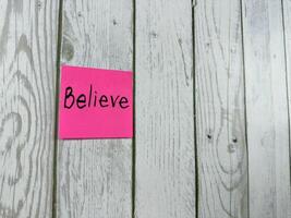 Pink sticky note with Believe written on it, placed on white wooden board. Ideal for conveying themes of belief, positivity and motivation. Perfect for concept ideas and personal growth photo