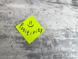 Bright yellow sticky note shaped as diamond featuring the word Positivity and smiley face on textured grey background, symbolizing cheerful mindset and the power of positive thinking in daily life photo