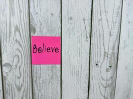 Pink sticky note with the word Believe written on it, pinned on rustic wooden board background. The image represents belief, positivity, and motivation. Ideal for concept themes photo