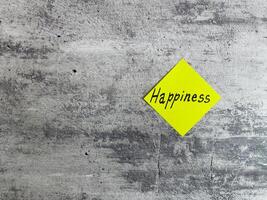 Bright yellow diamond-shaped sticky note with the word Happiness displayed on a textured grey background, representing joy, positivity, and the pursuit of a fulfilling life photo