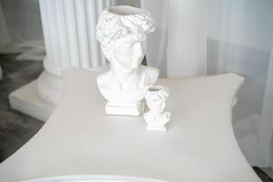 photo zone for photo sessions in antique and greek style antique sculptures in the interior. two plaster sculptures stand on the table. behind white chiffon fabric and columns