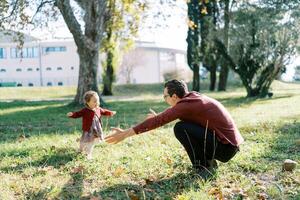 Little girl runs to her dad, who opened his arms in an embrace, squatting on the lawn photo