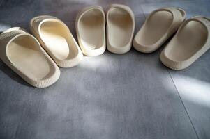 Three pairs of beige rubber slippers stand on a gray granite floor. A ray of sunlight falls on them. The slippers stand in a row, in a semicircle. They are the same size, viewed from above. Close-up photo