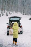 Woman stands near a car with an open trunk and a spread blanket inside in a winter park. Back view photo