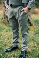 Groom with his hands in his pants pockets stands on the green grass. Cropped. Faceless photo