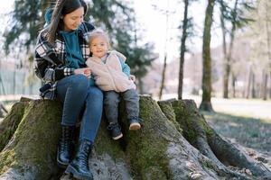 Smiling mom with a little girl are sitting hugging on a stump covered with green moss photo