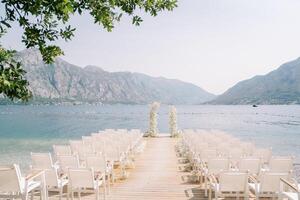 Rows of white chairs stand in front of a wedding arch on a pier by the sea against the backdrop of mountains photo