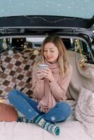 Smiling girl sits cross-legged on a blanket in the trunk of a car and looks at a cup of coffee in her hands photo