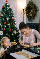 Little daughter looks at the cookies on the tray while mom rolls out the dough with a rolling pin photo
