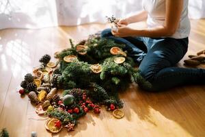 Girl with a branch of berries sits near a Christmas wreath on the floor. Cropped photo