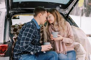 Man and woman touch foreheads while sitting with coffee mugs in car trunk in winter park photo