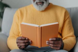 Senior man reads an orange book while seated comfortably on a sofa in a cozy living room during the afternoon photo