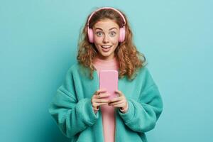 Young woman wearing pink headphones smiles while holding smartphone against a teal background in a bright setting photo