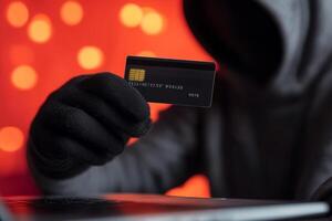 Individual in a hoodie holding a credit card in front of a laptop with a blurred orange background. Internet scammer hacker, Carding concept photo