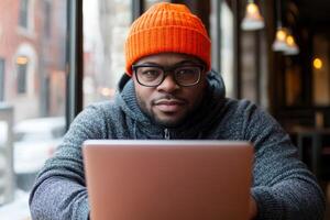 Man in an orange beanie works on a laptop while sitting by a window in a cozy urban cafe during winter photo