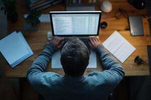 Person working late at a desk using a laptop surrounded by documents and a plant photo